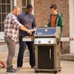 Three men standing next to a gas grill.