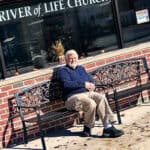 Man sitting on an outdoor bench in front of a building with windows displaying the text "driver of life choices.