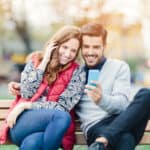 A smiling couple sitting on a bench while looking at a smartphone.