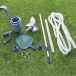 Swimming pool maintenance equipment displayed on grass, including a vacuum head, telescopic pole, skimmer net, and hoses.