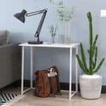 A minimalist workspace with a white desk featuring a black table lamp, a clear vase with greenery, and a brown backpack underneath, next to a potted cactus plant.