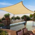 A patio with a beige sunshade covering, featuring wicker furniture, a pool with a waterfall feature, and potted plants.