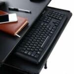 A desktop workspace featuring a black keyboard, a smartphone, a pen, and a leather-bound notebook.