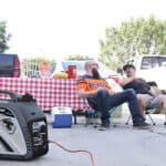 Two people sitting in folding chairs and chatting behind a pickup truck at a tailgate event, with a portable generator in the foreground.