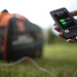 Charging a smartphone outdoors using a portable power source with a tent in the background.