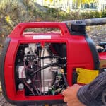 A person starting a portable red generator outdoors with autumn foliage in the background.