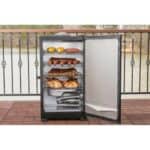 Outdoor electric smoker with multiple racks containing meats and vegetables on a balcony.