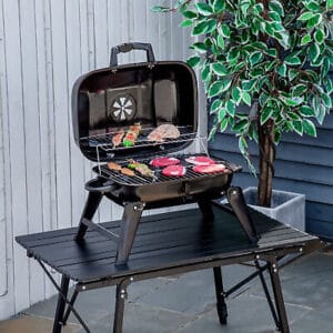Portable barbecue grill with various meats cooking on a table outdoors.