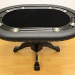 Oval poker table with black felt surface and padded edge, featuring built-in cup holders, on a wooden floor.