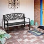 A black metal bench with decorative backrest on a tiled porch with a welcome mat and green cushions.