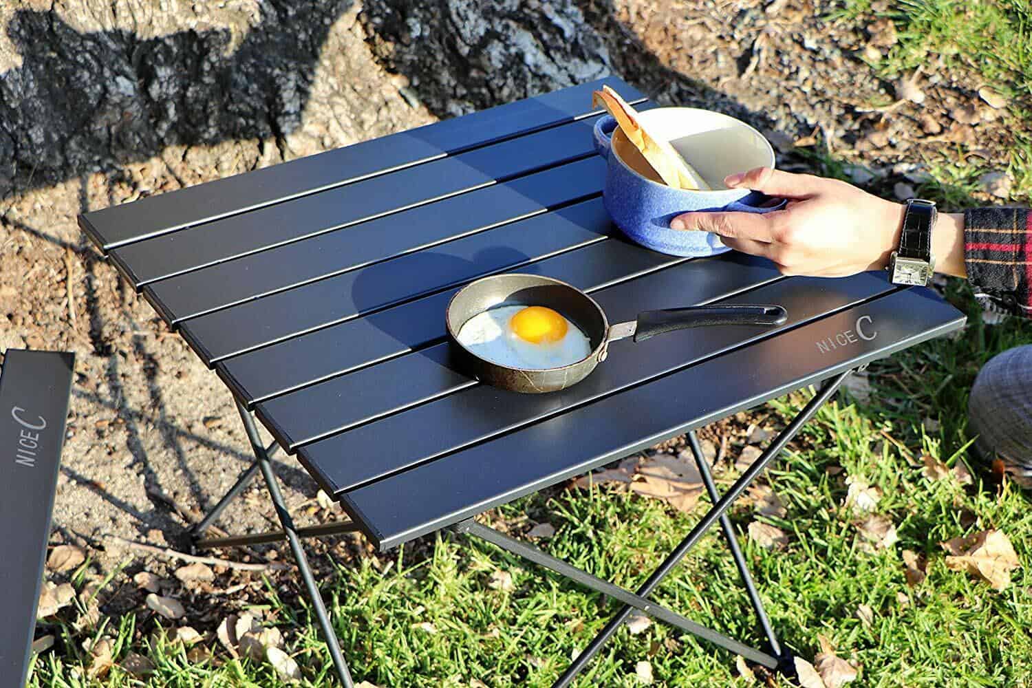 A person is holding a mug and frying an egg on a camping table.