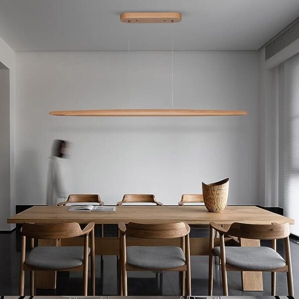 A minimalist dining room with a wooden table, chairs, and a 51" Wood Linear Pendant Light LED Dimmable Hanging Light Fixture. A blurred person is walking in the background.
