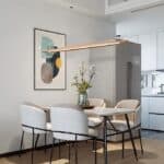 Modern kitchen with a round dining table, white chairs, and a 51" Wood Linear Pendant Light LED Dimmable Hanging Light Fixture above a gray refrigerator.