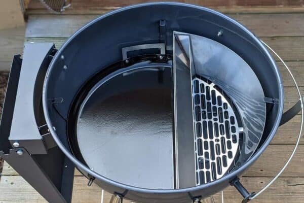 Top view of a round charcoal grill with an open lid, featuring a split Slow 'N Sear Drip 'N Roast Porcelain Grill Pan from SnS Grills on one side.