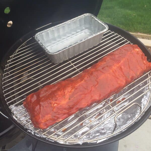 A large rack of ribs coated in barbecue sauce cooking on a charcoal grill, placed next to a Slow 'N Sear Stainless Steel Charcoal Basket for 18" Charcoal Grills from SnS Grills in an outdoor setting.