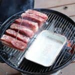 Ribs seasoned with spices are cooking on Slow 'N Sear Stainless Steel Charcoal Baskets for 18" Charcoal Grills, alongside a metal drip tray on a wooden deck.