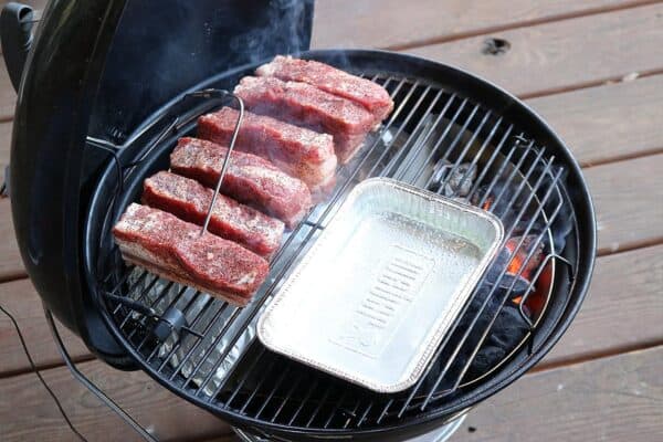 Ribs seasoned with spices are cooking on Slow 'N Sear Stainless Steel Charcoal Baskets for 18" Charcoal Grills, alongside a metal drip tray on a wooden deck.