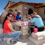 Group of seven people dining at a table made of salt blocks, with a rustic building in the background, in a bright, sunny salt flat setting. They are enjoying drinks chilled by The Ultimate Mini Ice Cube Maker Pink Silicone Bucket Ice Mold and Storage Bin Portable 2 in 1 Ice Cube Maker.