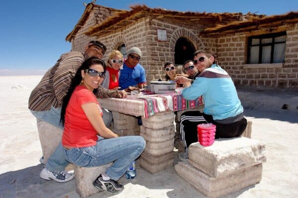 Group of seven people dining at a table made of salt blocks, with a rustic building in the background, in a bright, sunny salt flat setting. They are enjoying drinks chilled by The Ultimate Mini Ice Cube Maker Pink Silicone Bucket Ice Mold and Storage Bin Portable 2 in 1 Ice Cube Maker.