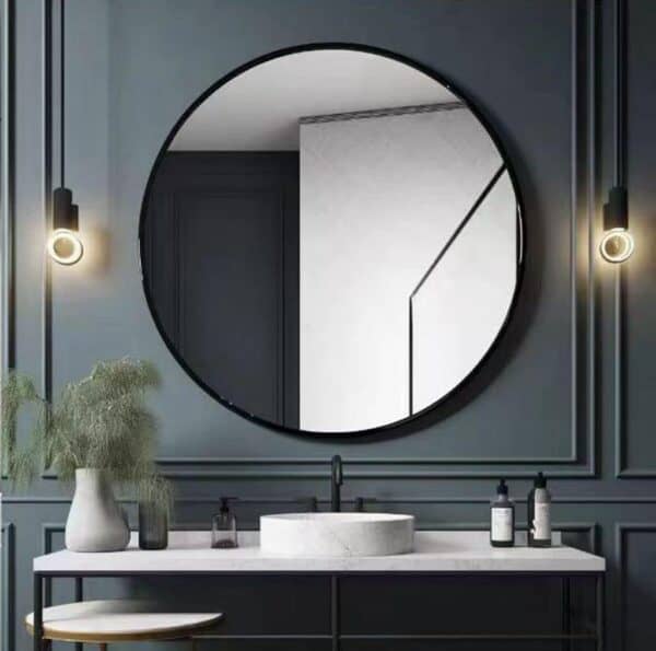 A modern bathroom featuring a 30 Inch Black Round Mirror Metal Frame mirror above a vanity with a vessel sink, flanked by two pendant lights, against a dark gray paneled wall.