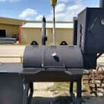 A large black Custom Made Heavy Duty BBQ Smoker Grill Steel on Casters Outdoor outside a business, featuring multiple compartments and a chimney, with a street and buildings in the background.