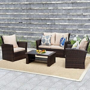 Outdoor patio Set of 4 Rattan Wicker Cushioned Furniture Set with two armchairs, a sofa, and a coffee table arranged on a rug against a gray brick wall, decorated with cushions and a fruit bowl on the table.