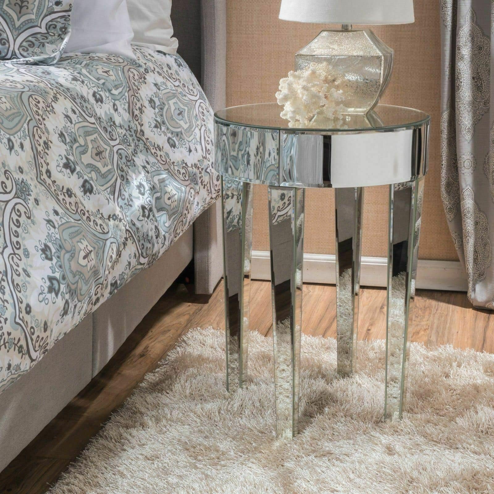 A modern bedroom featuring an Alvo Modern Glam Round Mirrored Side Table with Tapered Legs, a glass lamp, next to a bed with paisley-patterned bedding and set on a plush white rug.