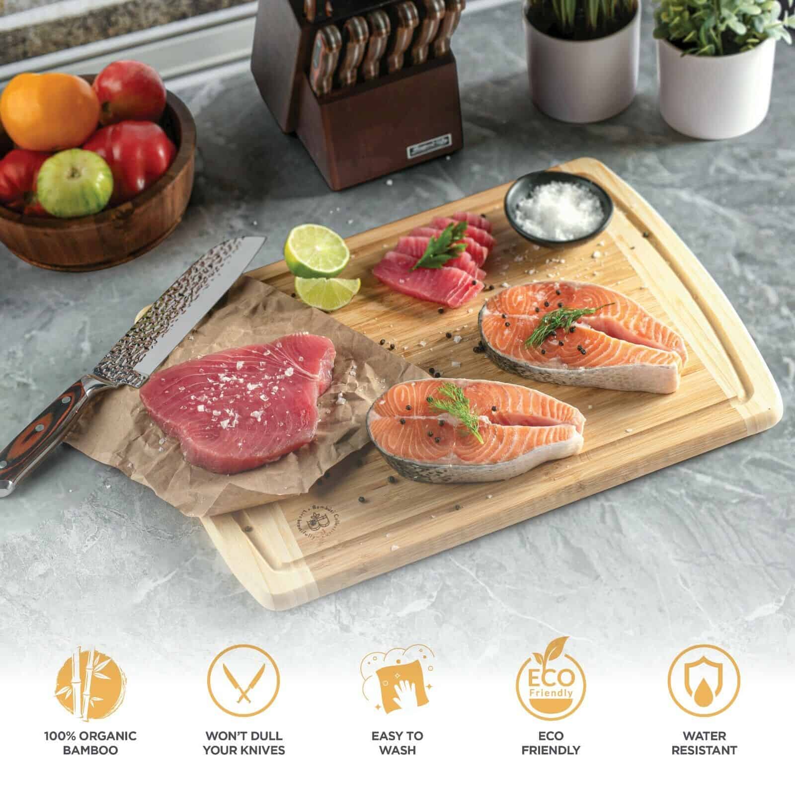 Sentence with product name included: Top view of a Premium Thick Bamboo Cutting Board Set of 2-Juice Grooves with raw salmon slices, a salmon steak, herbs, and lime on a kitchen countertop, with eco-friendly icons displayed below.